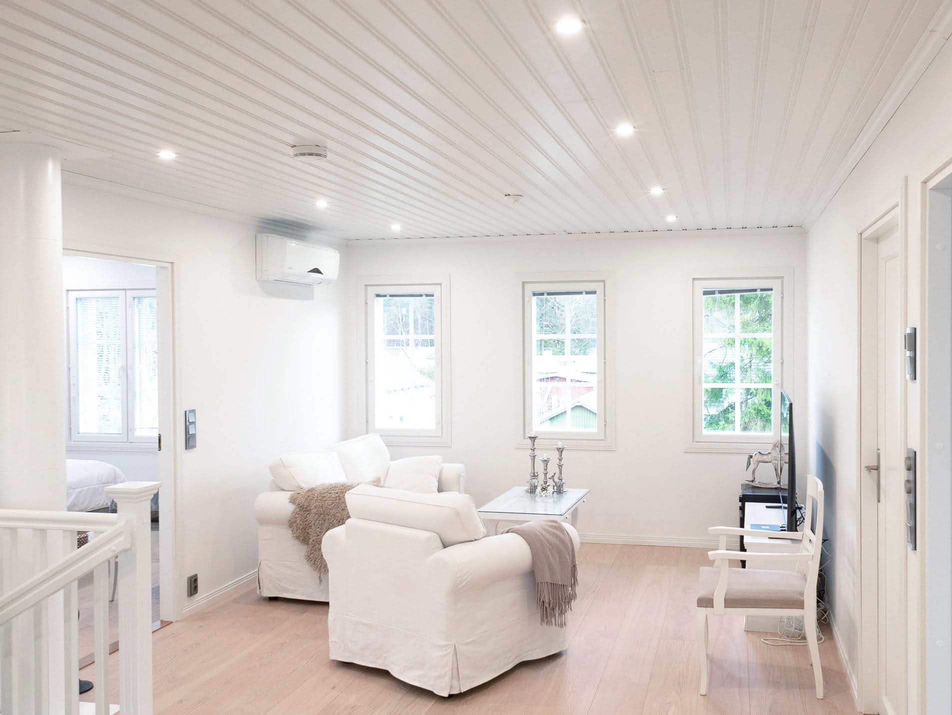 Moderni olohuone, jossa on LED-valaistus, valkoiset nojatuolit, sohvapöytä ja TV; lisää luonnonvaloa suurista ikkunoista ja korostaa puulattiaa.