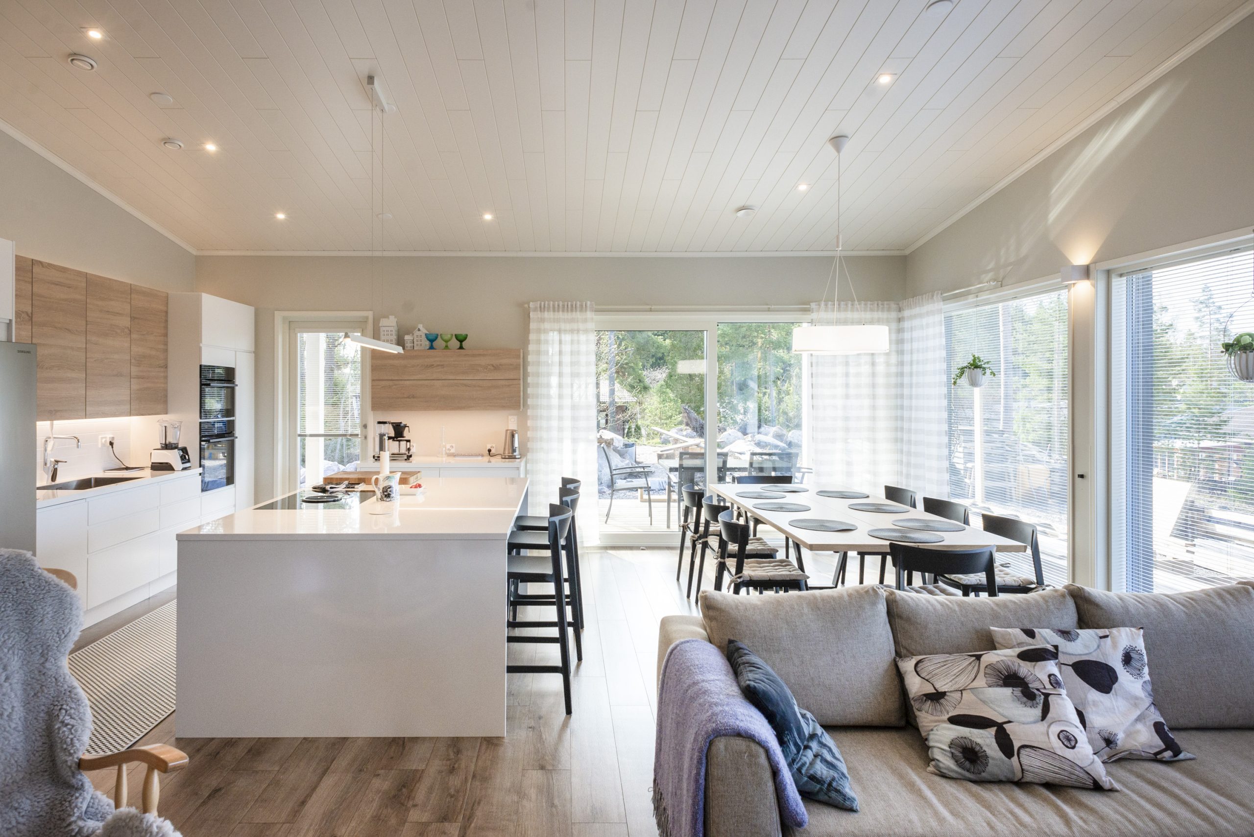 Valoisa ja moderni avokeittiö sekä olohuone, joissa valkoiset seinät ja katto. Tilaa hallitsee suuri saareke, jolla on baarituoleja, ruokapöytä mustilla tuoleilla ja mukava sohva. LED-valaistus täydentää suurista ikkunoista tulvivaa luonnonvaloa tarjoten näkymän vehreään ympäristöön. LedStore.fi:stä löydät laadukkaat LED-valaisimet juuri tähän tyyliin sopivana!