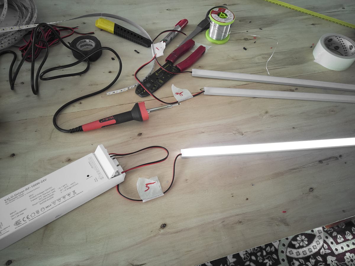 Työpöytä sähkötyökaluilla, LED-nauhat, johdot, loisteputket hehkuvat; sisältää pihdit, yleismittarin, juotoslangan ja virtalähteen.