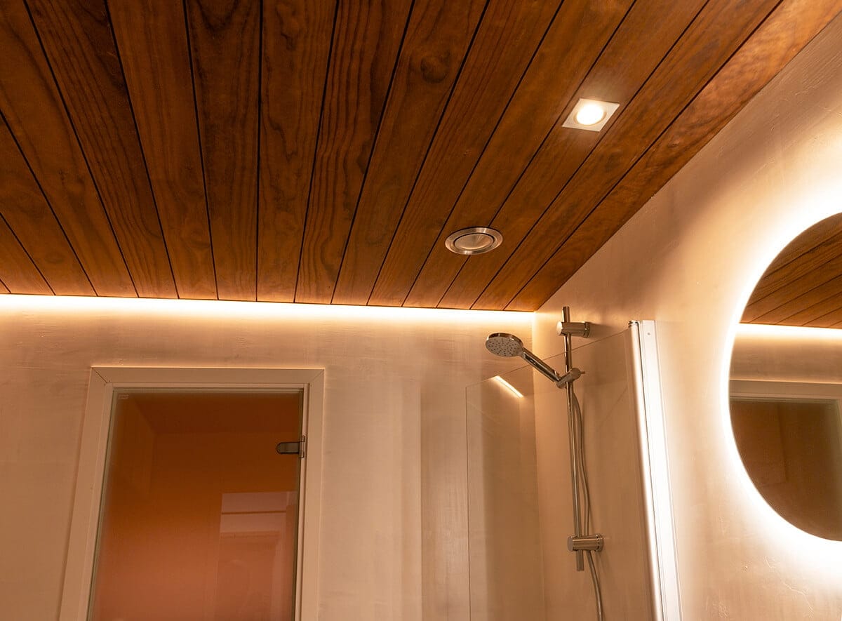 Puinen kattokylpyhuone, jossa ympäristön LED-valaistus, moderni suihkupää, kohdevalo ja pyöreä LED-peili; oviaukko vasemmalla.