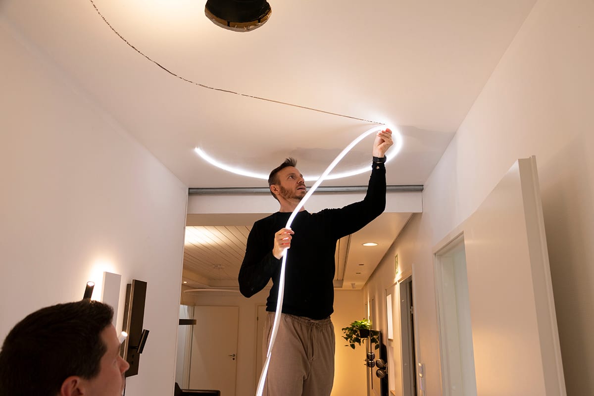 Henkilö kiinnittää joustavan LED-nauhavalaisimen moderniin kattoon, mikä parantaa huoneen valaistusta. Musta villapaita, beige housut; toinen henkilö etualalla, epätarkka.