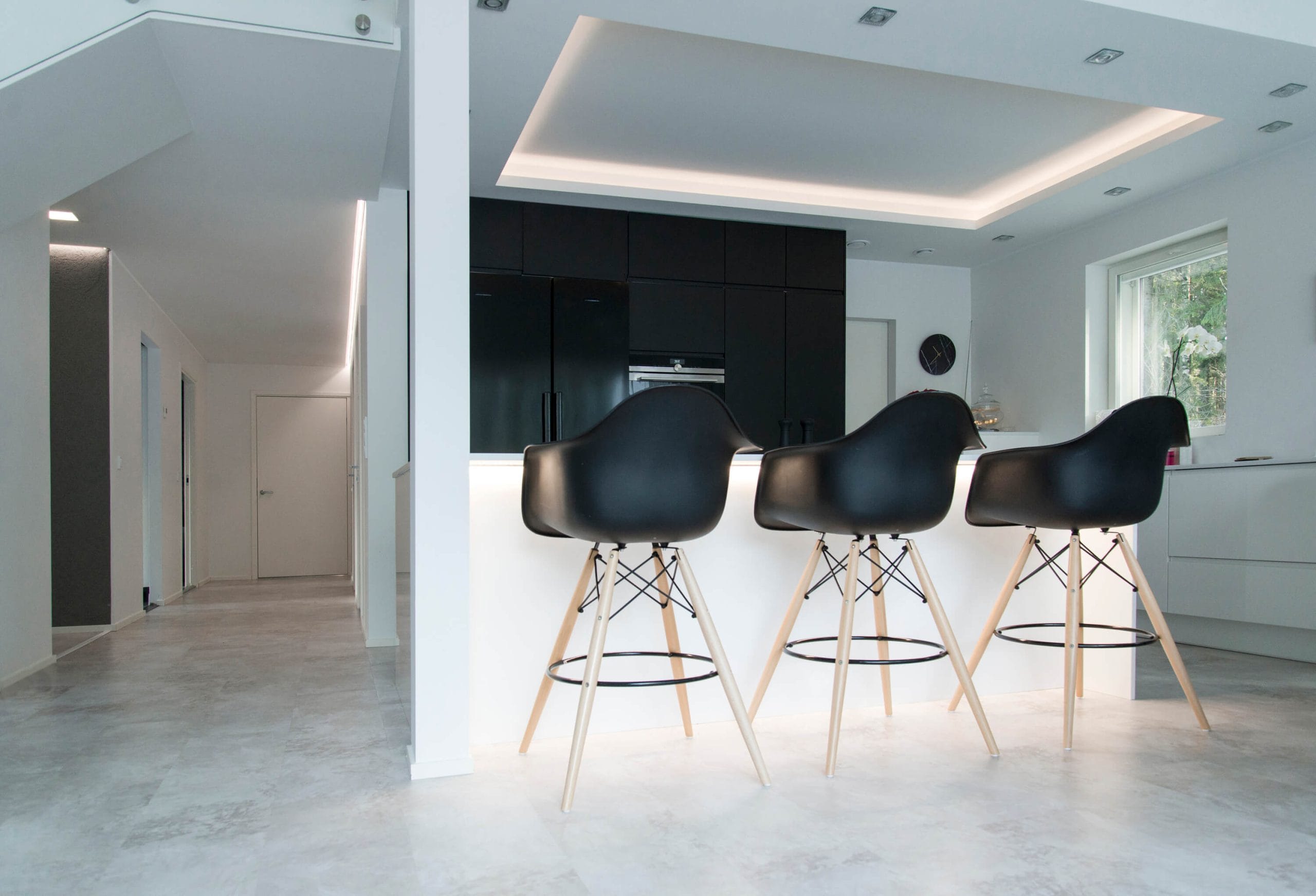 Moderni keittiö, jossa mustat baarituolit valkoisella saarella, tummat kaapit, upotettu katto LED-valaistuksella, ikkuna ja minimalistinen eteinen luovat tyylikkään ilmeen.