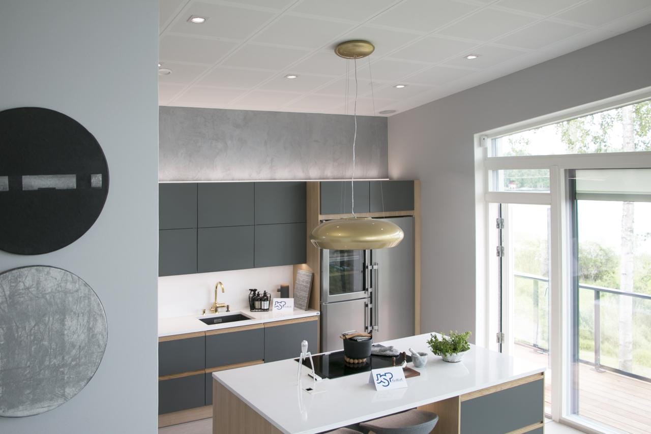 Moderni keittiö, jossa tummanvihreät kaapit, valkoinen saari, kultaiset riippuvalot, kodinkoneet; LED-korostettu seinätaide, joka korostaa luonnonvaloa suurista ikkunoista.