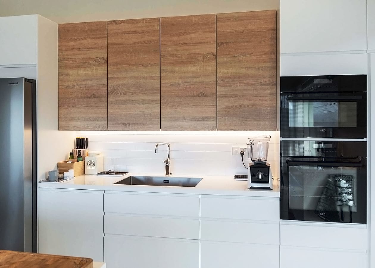 Moderni keittiö, jossa on puiset yläkaapit, LED-valaistut valkoiset työtasot, ruostumattomasta teräksestä valmistettu pesuallas kromihanalla, kaksinkertainen uuni ja työtasolaitteet.