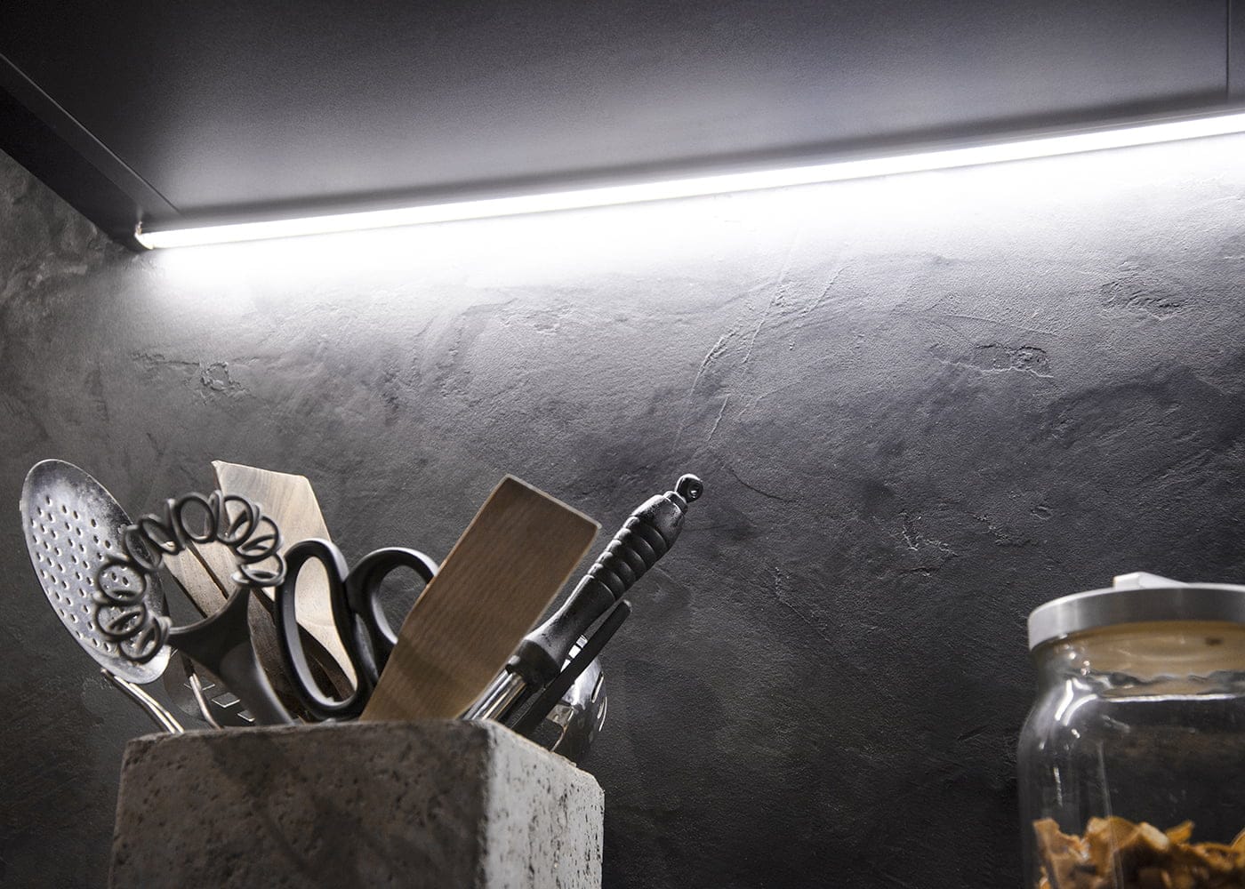 Keittiökuva, jossa astiat pidikkeessä, kirkas LED-nauhavalo korostaa kuvioitua tummaa seinää; lasipurkki näkyy oikealla. Ihanteellinen moderniin LED-valaistukseen.