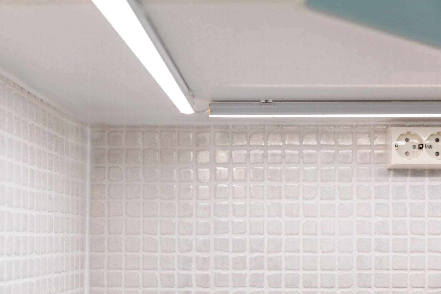 Valaistus_keittiö_6 - LedStore Moderni keittiön backsplash valkoisilla teksturoiduilla laatoilla, jotka valaisevat kaapin alla olevalla lineaarisella LED-valolla; lempeä hehku korostaa seinässä olevaa pistorasiaa.
