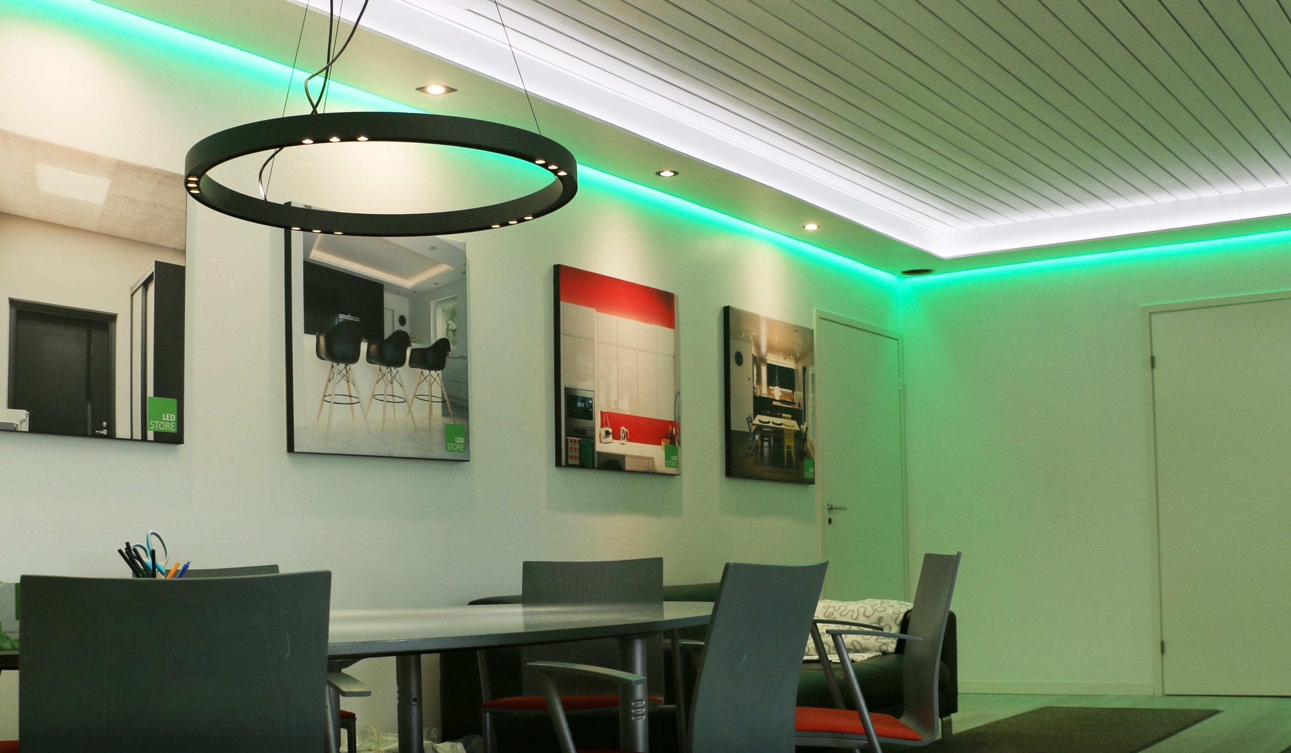Moderni huone, jossa on musta pyöreä LED-valo, harmaat tuolit pitkän pöydän ympärillä, kehystetty taide seinällä ja kattovalaistus vihreillä korosteilla tyylikkään tunnelman luomiseksi.