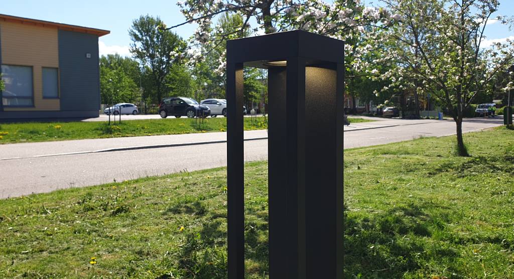 Moderni musta valaisinpylväs LED-valolla tien varrella; kukkivia puita, pysäköityjä autoja ja rakennus, jossa on suuret ikkunat taustalla aurinkoisen taivaan alla.