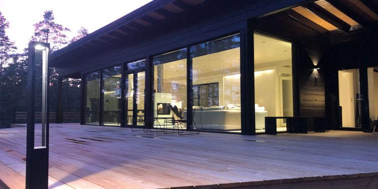 Moderni talo, jossa on suuret lasi-ikkunat ja tilava puinen terassi, ympäröivätynä puilla. Sisätilat ovat kirkkaasti valaistut ja esittelevät minimalistisen designin käsissä sävyissä. LedStore.fi:n LED-ulkovalot lisää lämpimän hehkun iltahämärään.