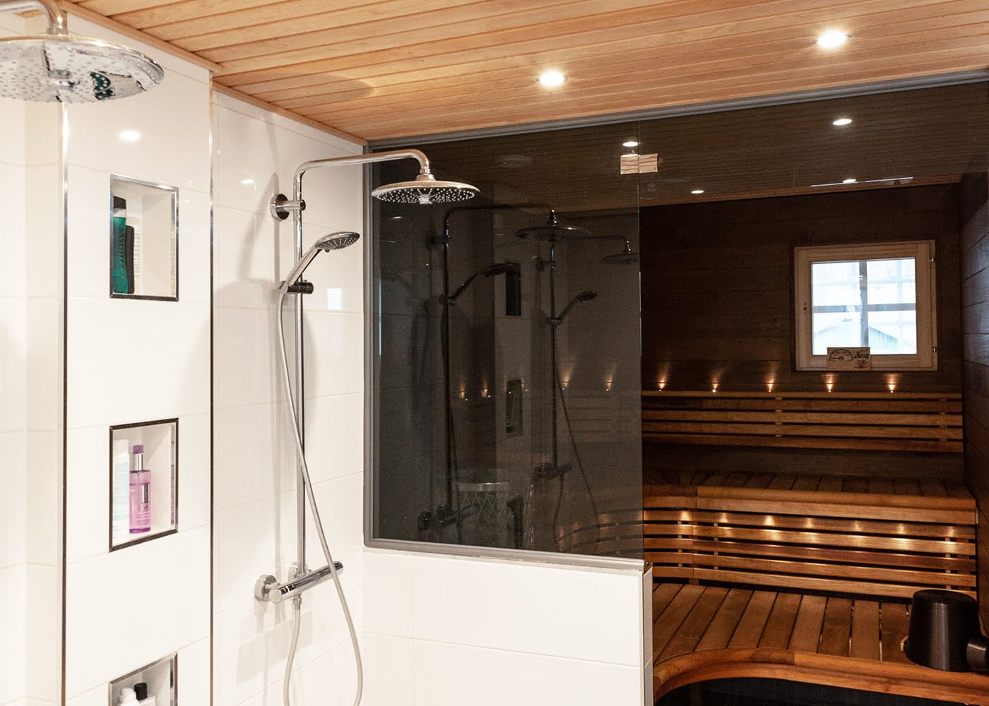 Moderni kylpyhuone kahdella sadesuihkulla, lasiseinä, sisäänrakennettu kylpytuotehylly, vieressä oleva puinen sauna, tunnelmallinen ikkunavalaistus ja LED-korostus.